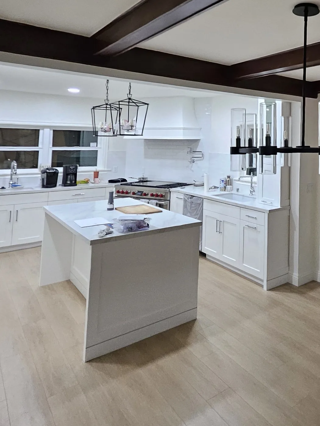 A finished kitchen open to a dining room, with white shaker cabinets, a stone island, black lantern pendants and stained ceiling beams
